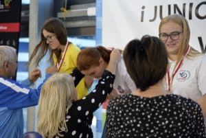 Kobieta stoi przed dziewczyną. Ta uśmiecha się do niej. Obok obieta nakłada medal na szyje dziewczyny. Ta pochyla się do przodu. Po lewej stronie mężczyzna patrzy na dziewczynę i ma ręce w geście już po założeniu medalu. Dziewczyna patrzy na niego.