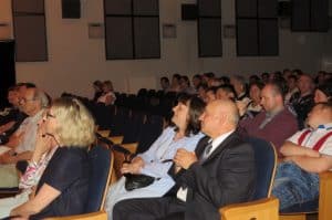 Ludzie siedzą na widowni. Ich twarze są zwrócone na wprost.