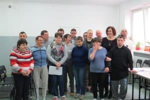 Grupa osób pozuje do zdjęcia. Są to kobiety i mężczyźni. Po lewej stronie na przedzie kobieta trzyma kartkę w dłoniach.