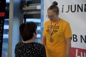 Kobieta stoi przed dziewczyną i patrzy na nią. Dziewczyna także. Ma na sobie medal.Kobieta stoi tyłem do kamery.