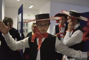 Mężczyzna w białej koszuli, czarnej kamizelce i kolorowym kapeluszem z dużym rondem pozuje do zdjęcia. Uśmiecha się.