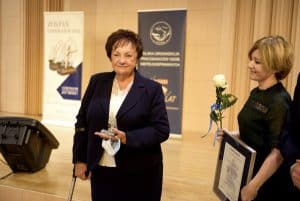 Kobieta z kulą stoi i trzyma statuetkę w dłoni. Pozuje do zdjęcia. Po prawej stronie stoi kobieta z dyplomem i różą. Jest bokiem do obiektywu.