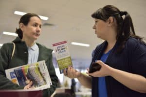 Dwie kobiety stoją obok siebie. Jedna ma twarz zwróconą ku drugiej. W dłoniach trzymają gazetki i ulotki.