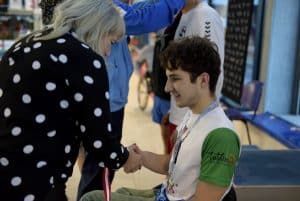 Kobieta gratuluje chłopcu przez podanie ręki. Chłopak uśmiecha się. Kobieta stoi tyłem do obiektywu a chłopak jest bokiem.