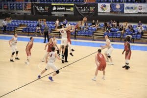 Mężczyźni grają na boisku. Na środku dwóch mężczyzn stykają się w powietrzu, w górze lecąca piłka. Wokół inni gracze.