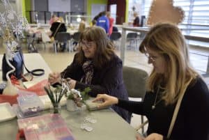 Kobieta siedzi przy style z rękodziełami. Obok niej kobieta w podeszłym wieku trzyma w dłoni wstążkę.