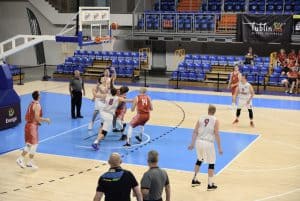 Mężczyźni są przy koszu od koszykówki. Pod koszem troje mężczyzn blisko siebie patrzących w górę na lecącą piłkę.