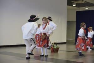 Ludzie w ludowych strojach są na środku sali. Ich twarze są zwrócone ku sobie.