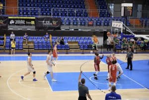 Sportowcy stoją pod koszem. Mężczyźni tuż pod koszem patrzą w górę, piłka jest w obrębie kosza.