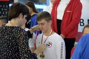 Kobieta stoi przed chłopcem. Ma on zamkniete oczy. KObieta jedną ręką poprawia medal.