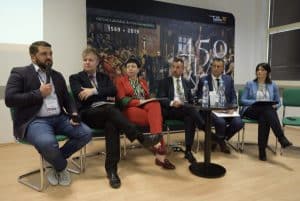 Grupa osób siedzi na środku sali. Są to czterej mężczyźni i dwie kobiety. Mężczyzna po lewej stronie trzyma mikrofon blisko ust. Za nimi plakat z obrazem “Unii Lubelskiej”.