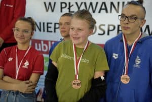 Po prawej stronie stoi dwóch młodych chłopców. Jeden z nich szeroko się uśmiecha. Po lewej stronie stoi uśmiechnięta dziewczyna.