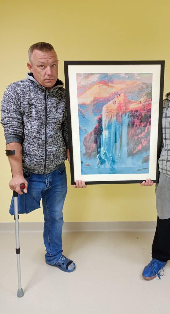 Mężczyzna stojący na jednej nodze posługujący się kulą trzyma w ręce duży obraz. Obok niego wycięty widok drugiego mężczyzny, który pomaga pierwszemu trzymać obraz.