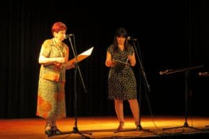 dwie kobiety stoją na scenie. Obioe trzymaja teczki w doniach. Prze dich ustami są stojące mikforony.