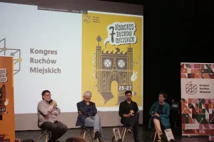 Na scenie siedzi czworo ludzi- trzech mężczyzn i kobieta. Mężczyzna pierwszy po lewej ma mikrofon i przemawia.