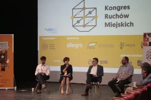 Na scenie siedzi czworo ludzi- trzech mężczyzn i kobieta. Kobieta przemawia do mikrofonu.