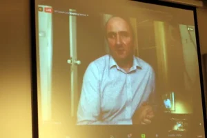 Młody mężczyzna stoi z mikrofonem i mówi do publiczności.