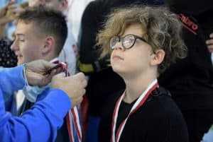 Młody chłopiec patrzy się na osobę stojącą przed nim. Ma na sobie medal. Osoba ma wyciągnięte ręce na wysokości jego twarzy i trzyma medal.
