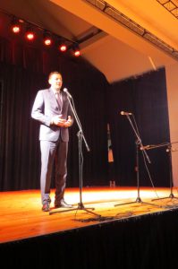 Mężczyzna stoi na scenie. Przed nim jest mirkfon stojący.