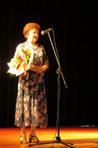 Kobieta stoi na scenie. Przed nią jest mirofon stojący.