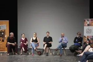 Na scenie siedzi sześć osób- dwie kobiety i czterech mężczyzn. Mężczyzna drugi od prawej przemawia do mikrofonu.