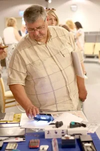 Starszy mężczyzna stoi nad stołem na którym znajdują się różne urządzenia. Trzyma w ręce jedno z nich i z zainteresowaniem ogląda.