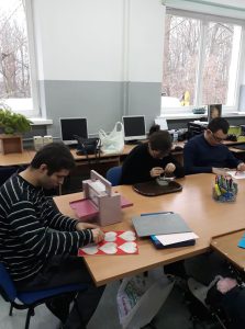 Trzech mężczyzn siedzi przy stołach. Jeden z nich ma przed sobą kartkę z serduszkami i trzyma na niej ręce. Dwójka innych siedzi pochyleni i wykonują czynności.