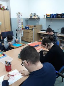 Mężczyźni siedzą przy stole. Jeden mężczyzna siedzi tyłem do obiektywu i trzyma kredkę czerwoną a przed sobą ma kartkę z rysunkiem. Po drugiej stronie na wózku inwalidzkim siedzi mężczyzna i dwoma palcami trzyma piłeczkę.