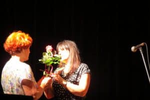Kobiety są na scenie. Jedna wręcza kwiaty drugiej.