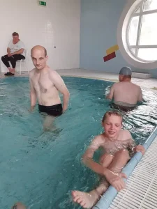 Mężczyźni kąpią się w basenie. Chłopiec mocno trzyma się końca basenu.