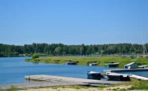 Krajobraz nad jeziorem. Na pierwszym planie znajduje się molo, przy nim przycumowane są trzy łódki. W oddali widać kolejne molo i cztery inne łódki przy brzegu. W oddali za wodą widać gęsty las.