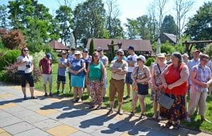 Grupa osób stoi na tle drzew oraz drewnianych budynków. Kobieta znajdująca się najbardziej po lewej stronie trzyma zawieszony na szyi aparat. Nad nimi znajduje się bezchmurne niebo.