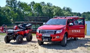 Dwa pojazdy strażackie stoją na plaży. Jeden z nich to quad a drugi to terenowe auto z napisem „STRAŻ” na otwartych drzwiach. Oba pojazdy są koloru czerwonego. W tle widać drewniany budynek, wodę oraz las.