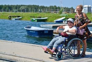 Dwie osoby stoją na molo nad wodą. Pani po lewej trzyma za rączki wózek, na którym siedzi druga kobieta. W tle znajdują się woda, łódki, pas zieleni oraz w oddali las.