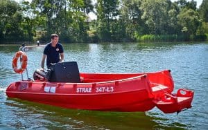 Na łodzi z napisem „STRAŻ 344-Ł3” stoi mężczyzna. Łódka znajduje się na wodzie, z tyłu przymocowane ma koło ratunkowe. W tle widać drzewa.
