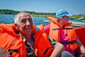 Dwie osoby w kapokach – od lewej mężczyzna oraz kobieta w czapce z daszkiem siedzą. W tle znajduje się woda, molo, pas zieleni oraz las.