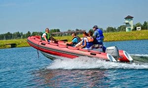 Grupa osób w kapokach znajduje się na pontonie, który widoczny jest z profilu. Z tyłu pontonu na wodzie widać pianę wodną. Jedna z osób siedzi na czubku pontonu, jest on lekko uniesiony z przodu. W tle widać pas zieleni oraz drzewa.