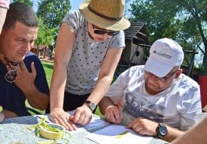 Trzy osoby znajdują się przy stole. Dwóch mężczyzn siedzi, jeden z nich po prawej ma przed sobą kartkę i dotyka jej długopisem. Kobieta na środku stoi opierając ręce o stół. W tle widać drewniany domek i drzewa.