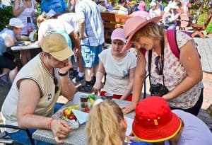 Grupa osób znajduje się przy stole. Cztery osoby – w tym dwie tyłem do obiektywu – siedzą a jedna kobieta z aparatem na szyi stoi nad nimi. Głowy mają skierowane w jednym kierunku. W tle znajdują się inne osoby, stoły oraz ławki.