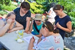 Pięć osób znajduje się przy stole. Dwie kobiety i mężczyzna siedzą, jedna z nich – siedząca po prawej stronie – trzyma w ręce długopis skierowany ku kartce papieru. Dwie inne kobiety stoją nad stołem, jedna z nich – po lewej – opiera się o niego ręką.