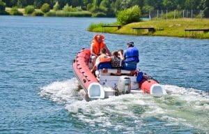 Grupa osób w kapokach znajduje się na pontonie, który odwrócony jest tyłem do obiektywu. Z tyłu pontonu na wodzie widać pianę wodną. Jedna z osób siedzi na czubku pontonu, jest on lekko uniesiony z przodu. W tle widać pas zieleni oraz drzewa.