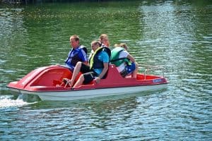 Na rowerku wodnym znajdującym się na wodzie siedzą cztery osoby w kapokach. Dwóch mężczyzn siedzi z przodu, a dwie osoby siedzą tyłem do nich.
