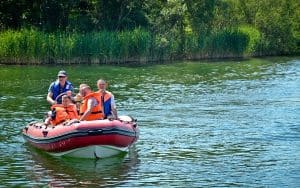 Na pontonie znajduje się grupa ludzi w kapokach. Ponton płynie po wodzie. W tle widać zielony brzeg porośnięty gęsto roślinnością oraz drzewa.