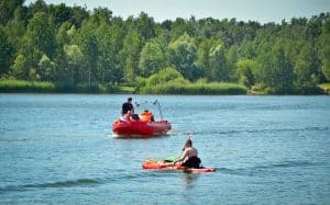 Na wodzie znajduje się ponton oraz osoba siedząca na desce z wiosłem w obu rękach. W tle znajduje się gęsto porośnięty brzeg oraz las.