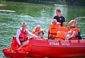 Na łódce znajduje się sześć osób, pięć z nich ma kapok. Jedna z osób – najbardziej po prawej stronie jest osobą poruszającą się na wózku. W tle widać wodę.