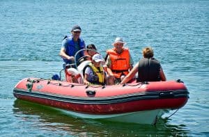 Na pontonie znajduje się pięć osób w kapokach. Jedna z nich – siedząca na czubku jest odwrócona tyłem do obiektywu. Dookoła nich znajduje się woda.