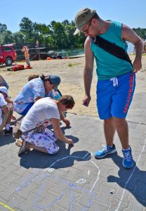 Na pierwszym planie na kostce brukowej znajduje się rysunek – głowa postaci z niebieską koronką. Po prawej stronie stoi mężczyzna, w prawej ręce trzyma kredę. Po lewej stronie kuca grupa czterech osób, widzimy je profilem do obiektywu, najlepiej widoczne są dwie pierwsze osoby. W tle znajduje się jezioro, piasek oraz drzewa.