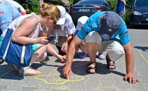 Na pierwszym planie kuca mężczyzna oraz kobieta. Mężczyzna w prawej ręce trzyma żółtą kredę – dotyka ona kostki brukowej, na której jest rysunek postaci. Kobieta po lewej palcem lewej ręki wykonuje gest wskazywania w kierunku jakiejś części rysunku, a w prawej ręce trzyma telefon.