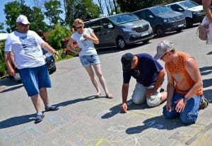 Na kostce brukowej znajdują się rysunki namalowane kredą. Po prawej stronie dwóch mężczyzn klęczy na kolanach, obaj mają w prawych rękach kredę. Po prawej stoją kobieta i mężczyzna – oboje mają głowy skierowane w kierunku rysunków.