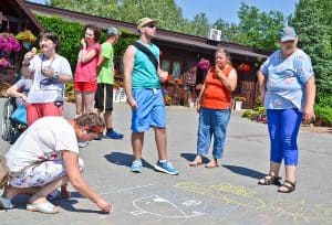 Na kostce brukowej znajdują się rysunki namalowane kredą. Po lewej stronie kuca kobieta, trzyma w prawej ręce kredę. Po prawej dwie koebiety i mężczyzna. W tle widać inne osoby oraz drewniany budynek a za nim drzewa.